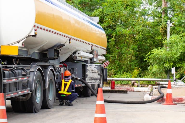 A large fuel tanker truck connected to a hose, actively refueling an external storage tank or facility.