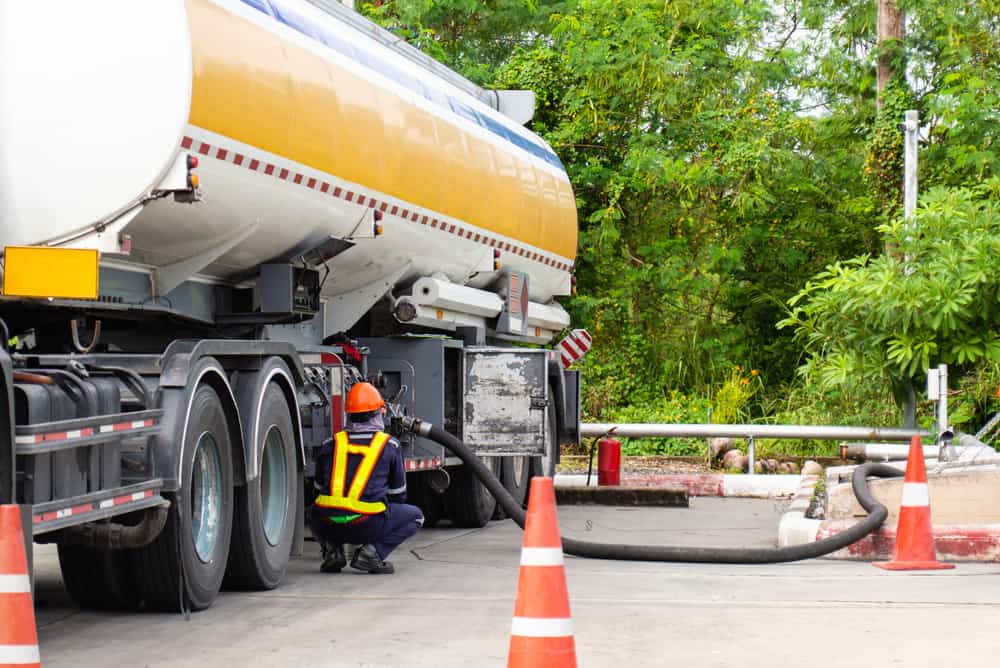 A close-up of a large yellow-and-white tanker truck alongside traffic cones, with a worker in protective clothing operating a fuel hose to refuel another vehicle or equipment.