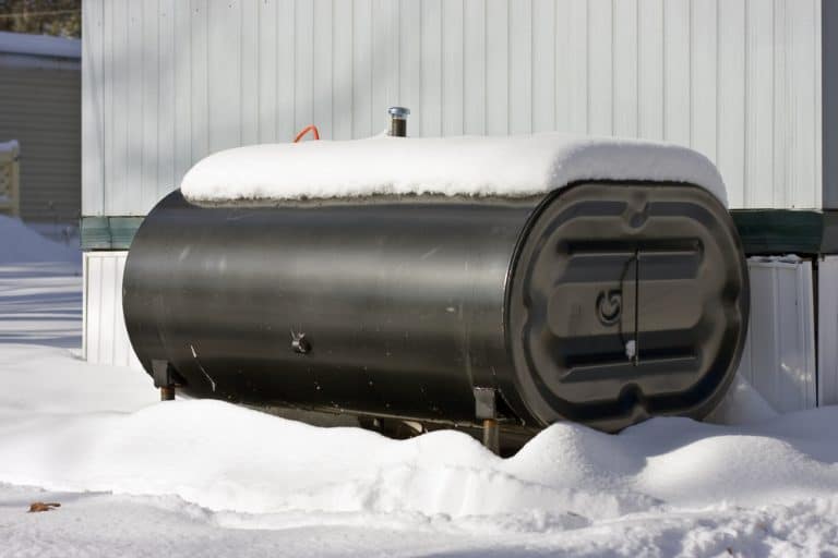 A large black horizontal heating oil tank sits outdoors against a light-colored exterior wall, its top and surrounding ground capped with a thick layer of snow.