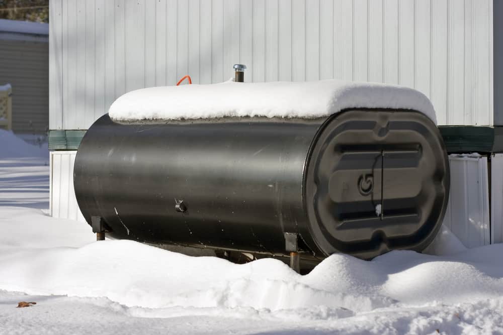 A large black horizontal heating oil tank sits outdoors against a light-colored exterior wall, its top and surrounding ground capped with a thick layer of snow.