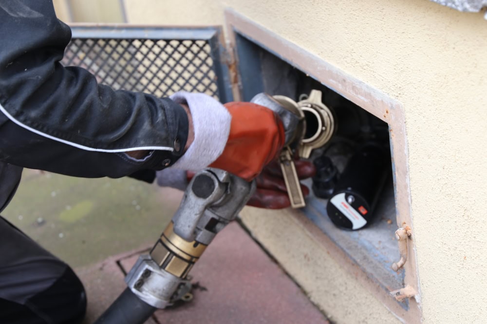 A heating oil delivery truck parked outside a home, with a worker connecting a hose to fill the home’s outdoor oil storage tank for the heating system.
