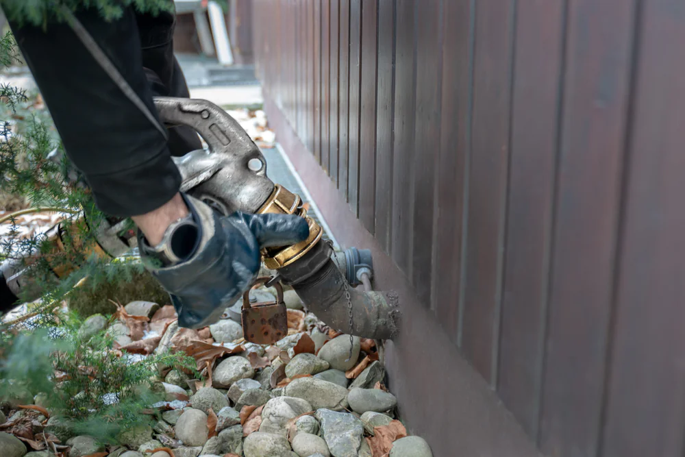 A person wearing gloves connects a metal hose to an outdoor pipe on the side of a building, surrounded by rocks and a small tree.