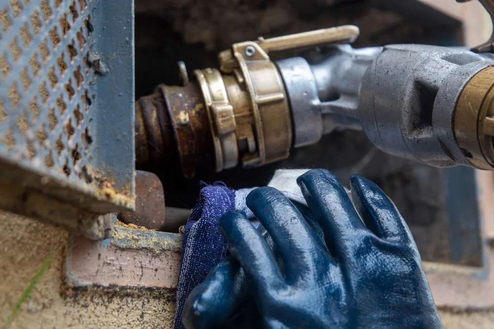 A person wearing blue protective gloves operates a metal hose connection attached to an industrial pipe opening on a concrete wall.