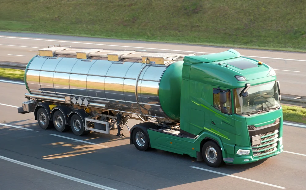 A green tanker truck with a shiny, silver tank is driving on a highway with grass along the roadside, heading in the left direction.