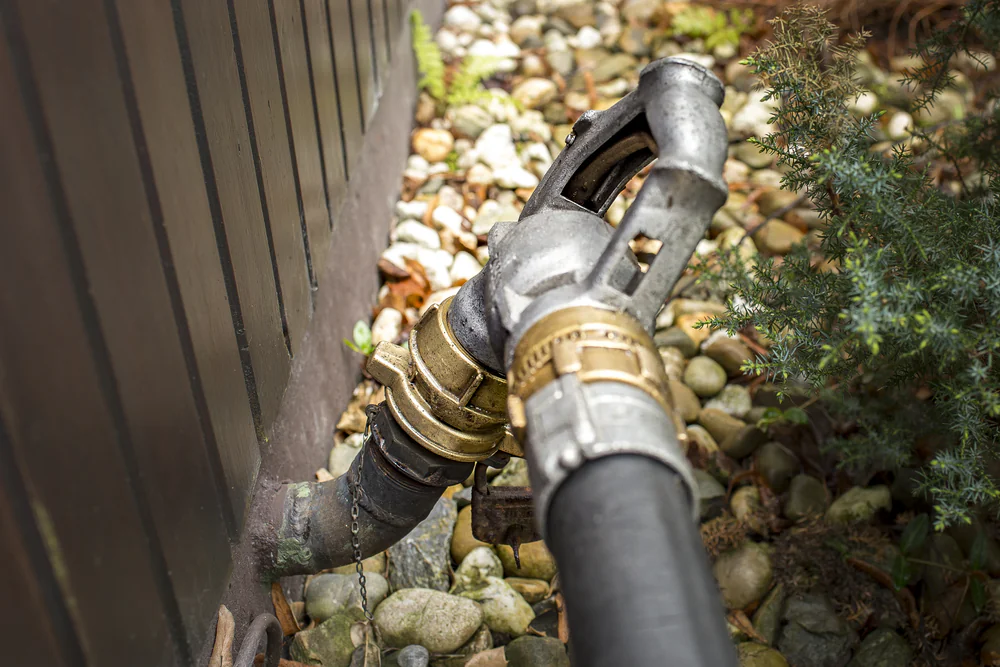 A close-up of a metal and rubber pipe connected to a wall outdoors, surrounded by small rocks and green plants.