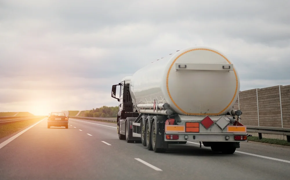 A white tanker truck drives on a highway, following a car, with the sun setting in the background and a sound barrier wall along the road.