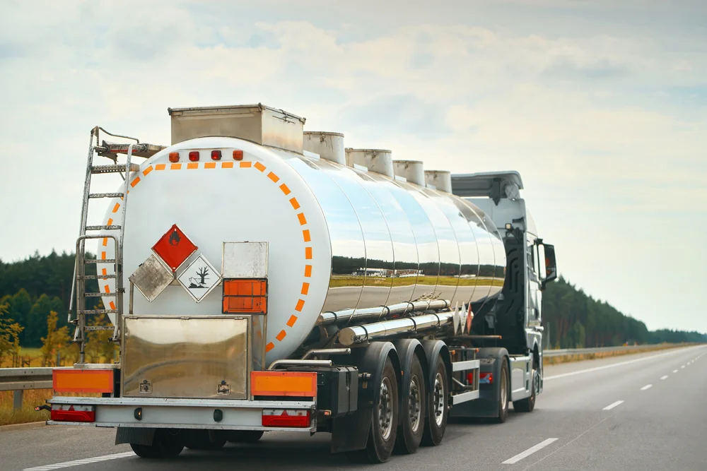 A large silver fuel tanker truck drives on an empty highway, with warning signs indicating hazardous materials. Trees line the road under a cloudy sky.