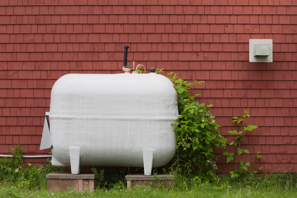 A large white propane tank stands on concrete blocks in front of a red shingle wall, surrounded by green grass and leafy plants.