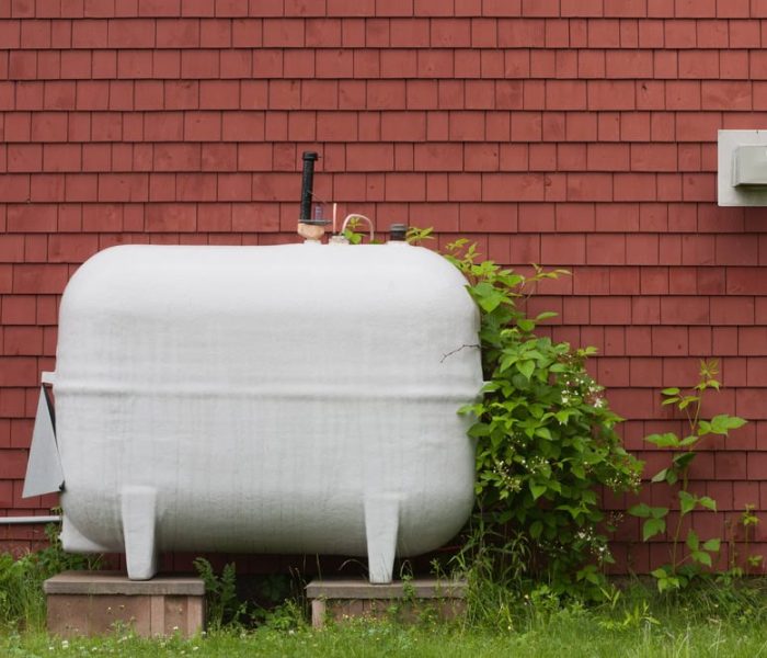 A large cylindrical heating oil tank placed outdoors directly beside the exterior wall of a building, with metal pipes and fittings for supplying fuel to the home’s heating system.