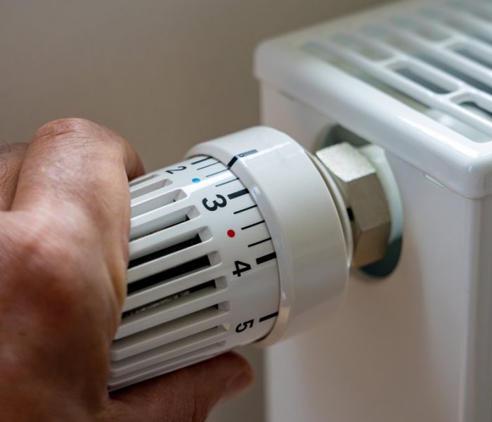 A man standing indoors adjusting a wall-mounted thermostat, lowering the temperature setting to reduce heating costs during an energy crisis.