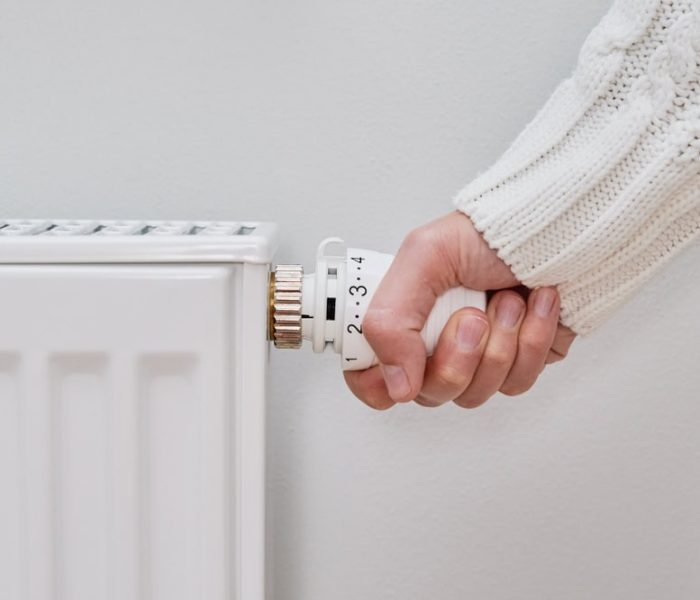 A woman crouching beside a home heating radiator, turning the valve to adjust the temperature as part of conserving energy during a crisis.