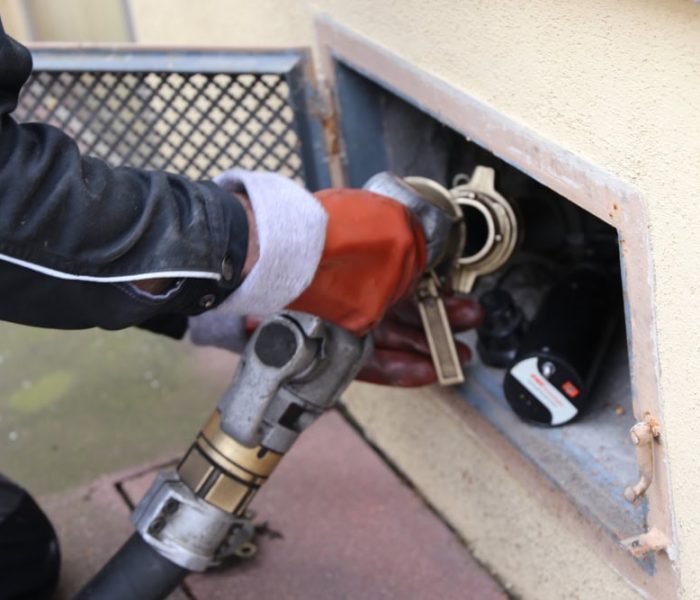 A heating oil delivery truck parked outside a home, with a worker connecting a hose to fill the home’s outdoor oil storage tank for the heating system.